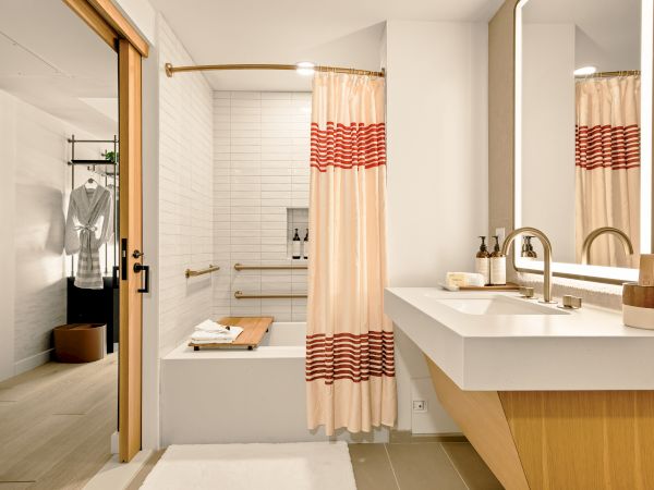 A modern bathroom with a bathtub, peach shower curtain, white sink, and a wooden vanity; soft lighting and a doorway leading to a wardrobe area.