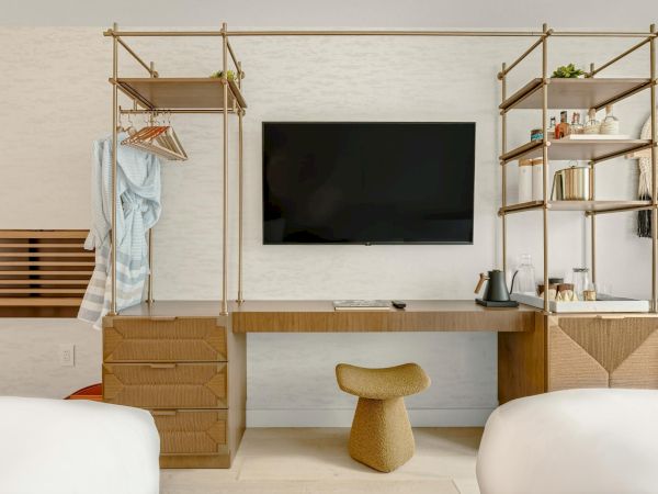 A stylish bedroom workspace with a mounted TV, modular gold metal shelves, a long wooden desk, and a small cushioned stool between two beds.