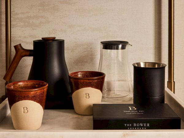 A stylish coffee setup with a black kettle, glass carafe, two ceramic cups labeled &ldquo;B,&rdquo; a small beige cup, and a sleek black box on a tray.