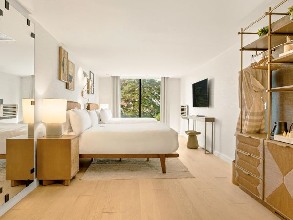 A bright, modern hotel-like bedroom with a large bed, wardrobe mirrors, a small desk, TV, and open shelving filled with decor accessories, all bathed in natural light.