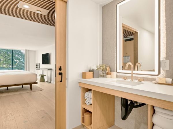 A chic hotel-style bathroom with a large mirror and wooden vanity, opening into a bright bedroom with a bed and seating, inside a modern suite.