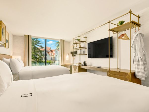 A bright hotel room with two white beds, a wall-mounted TV, open shelving, a robe on a stand, and a window view of trees and buildings, inviting and tidy.