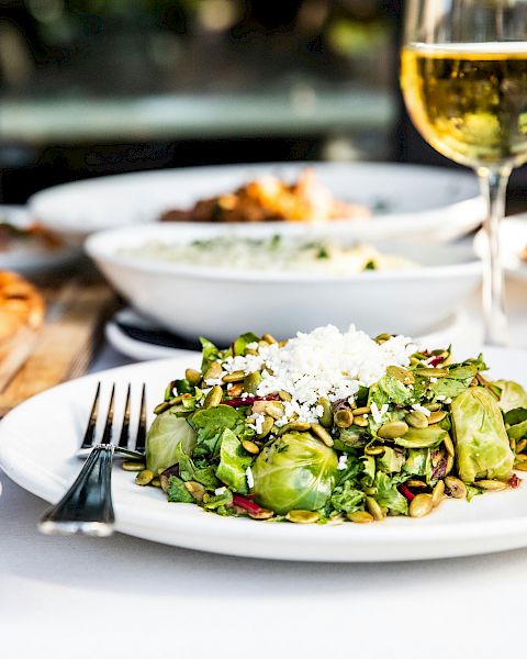 A table spread with a fresh green salad topped with seeds and cheese, alongside other dishes and a glass of white wine.