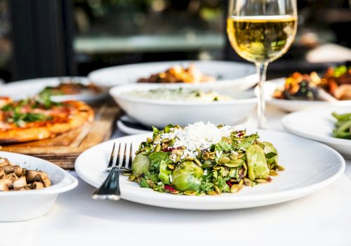 A table spread with a fresh green salad topped with seeds and cheese, alongside other dishes and a glass of white wine.