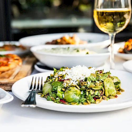 A table spread with a fresh green salad topped with seeds and cheese, alongside other dishes and a glass of white wine.