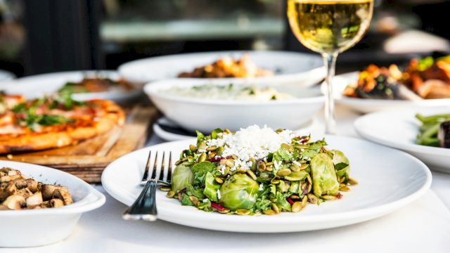 A table spread with a fresh green salad topped with seeds and cheese, alongside other dishes and a glass of white wine.