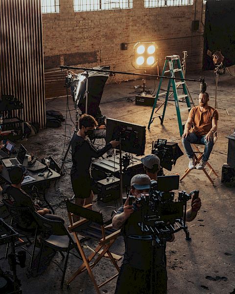 A film/TV crew shoots a scene in a warehouse studio with cameras, monitors, and bright stage lights.
