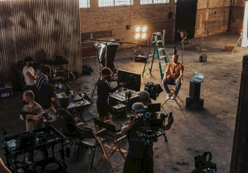 A film/TV crew shoots a scene in a warehouse studio with cameras, monitors, and bright stage lights.