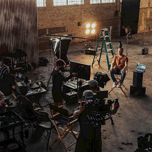 A film/TV crew shoots a scene in a warehouse studio with cameras, monitors, and bright stage lights.