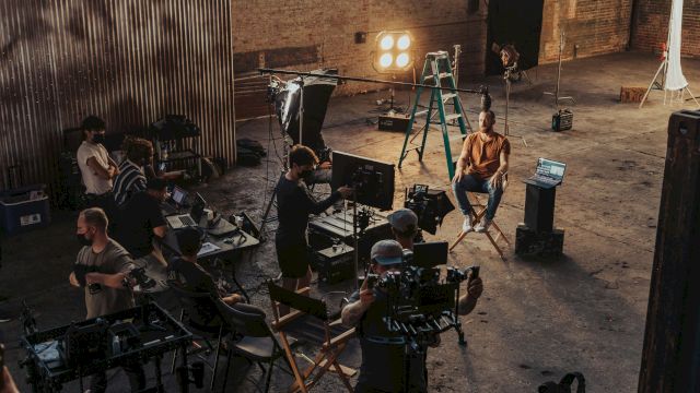 A film/TV crew shoots a scene in a warehouse studio with cameras, monitors, and bright stage lights.