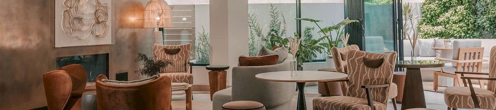 A modern lounge with upholstered chairs and sofas, a central table, lots of plants, and a ceiling covered in decorative flower-like lights.