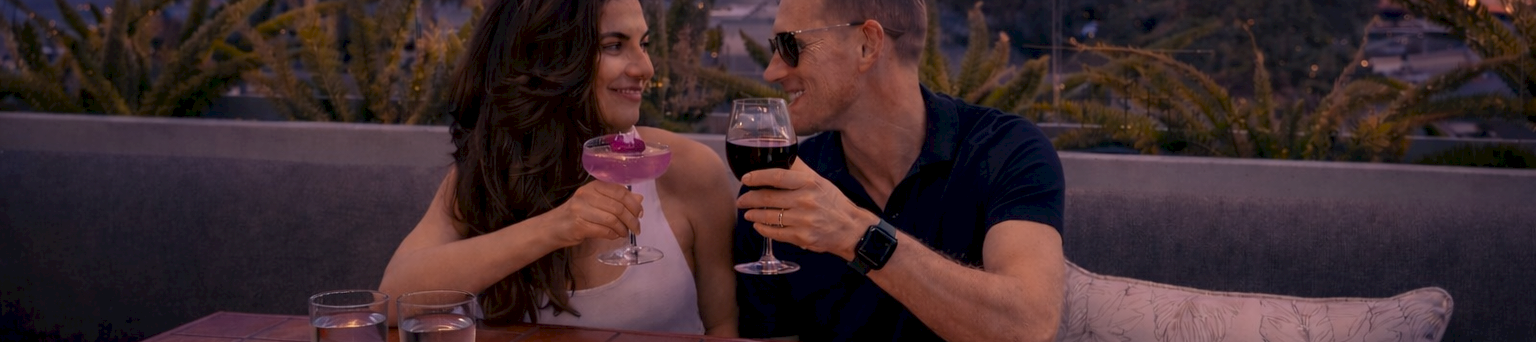 A couple cheers on a rooftop patio at sunset, holding wine glasses beside a candle and a rose in warm city lights.