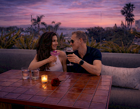 A couple cheers on a rooftop patio at sunset, holding wine glasses beside a candle and a rose in warm city lights.