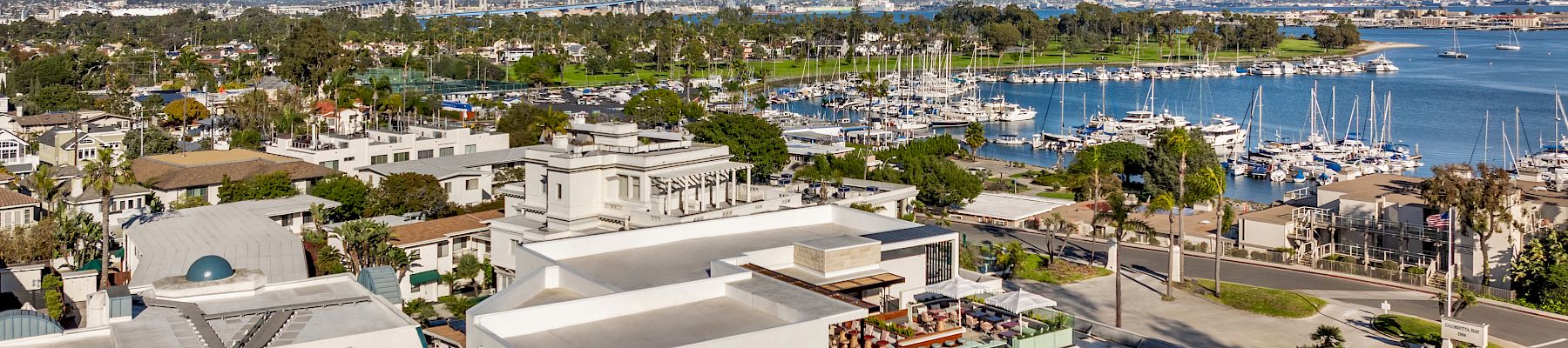 A sunny seaside view with the &ldquo;The Bower&rdquo; hotel, a marina packed with sailboats, and blue ocean and hills beyond.
