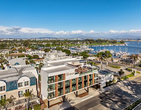 A sunny seaside view with the “The Bower” hotel, a marina packed with sailboats, and blue ocean and hills beyond.