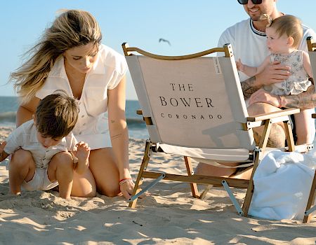 A sunny beach family lounges on sand while parents and kids sit by a “The Bower Coronado” chair, smiling.