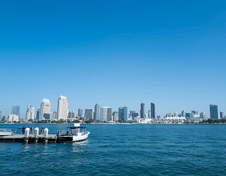 A sunny waterfront skyline with tall buildings, a dock, and small boats on the water.