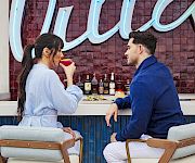 A couple enjoys drinks at a bar, facing each other beneath bright blue neon letters.