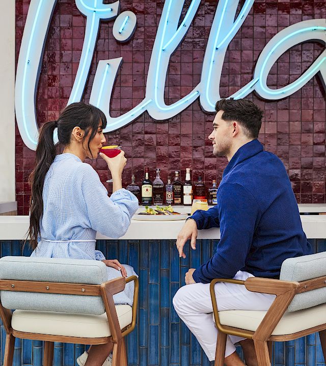 A couple enjoys drinks at a bar, facing each other beneath bright blue neon letters.