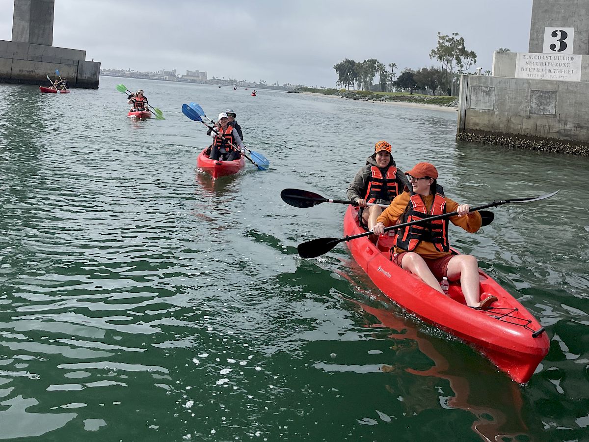 Kayakers in orange life vests paddle under a large bridge along the river near a U.S. Coast Guard checkpoint sign.