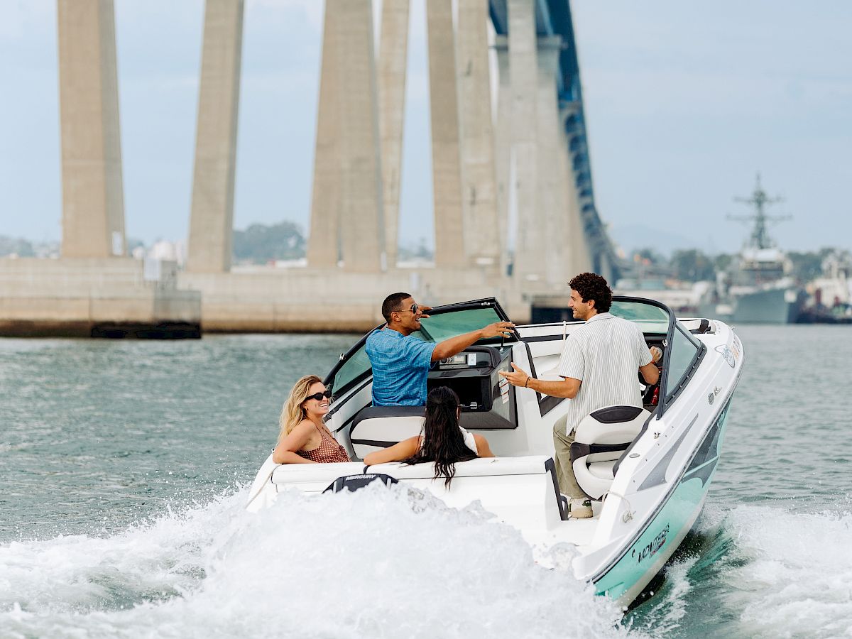 A speedboat races under a large bridge while friends enjoy a sunny ride, splashing in the water.