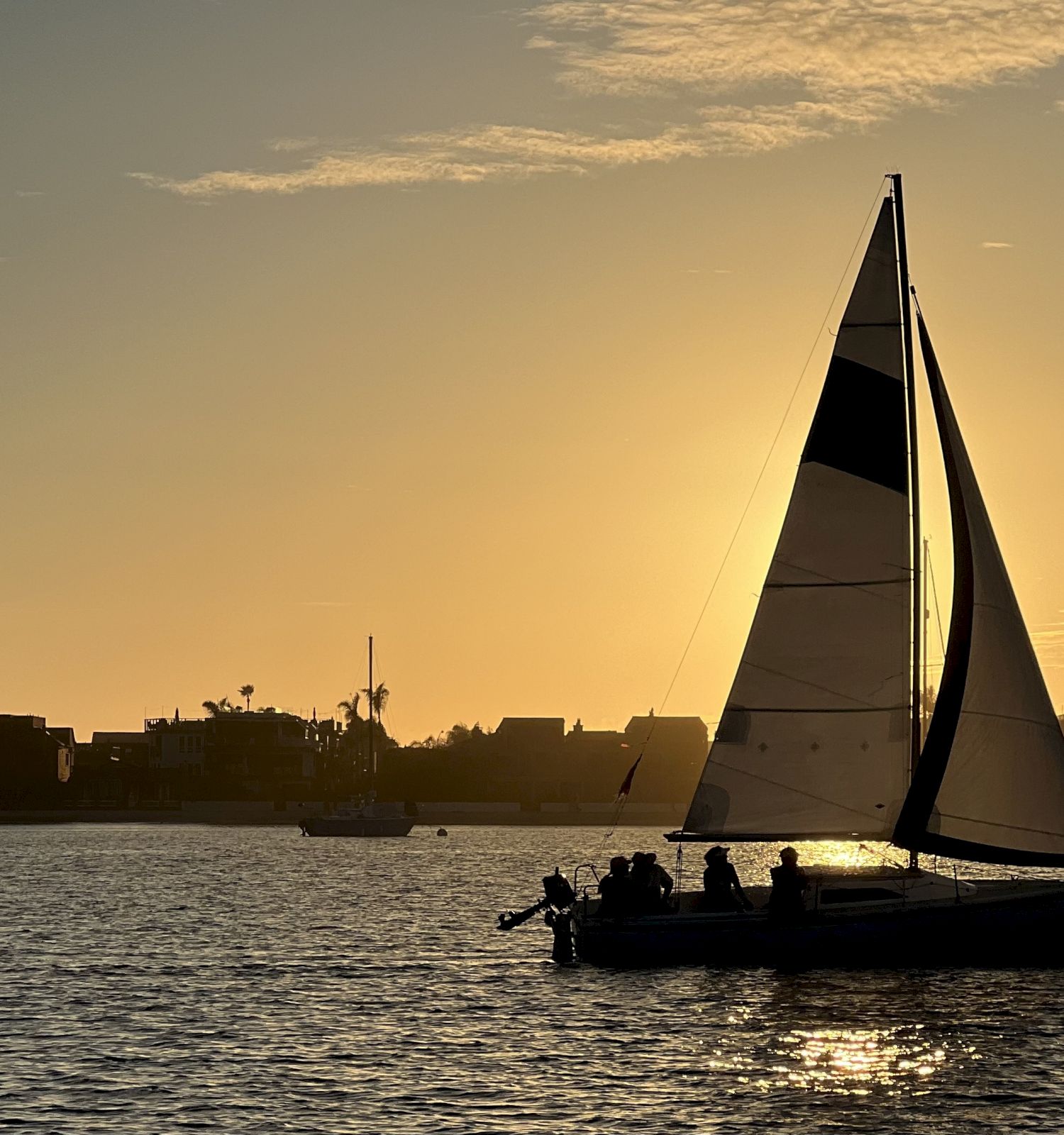 A sailboat glides across calm water at sunset, its sails catching the golden sky above a distant shoreline of buildings.