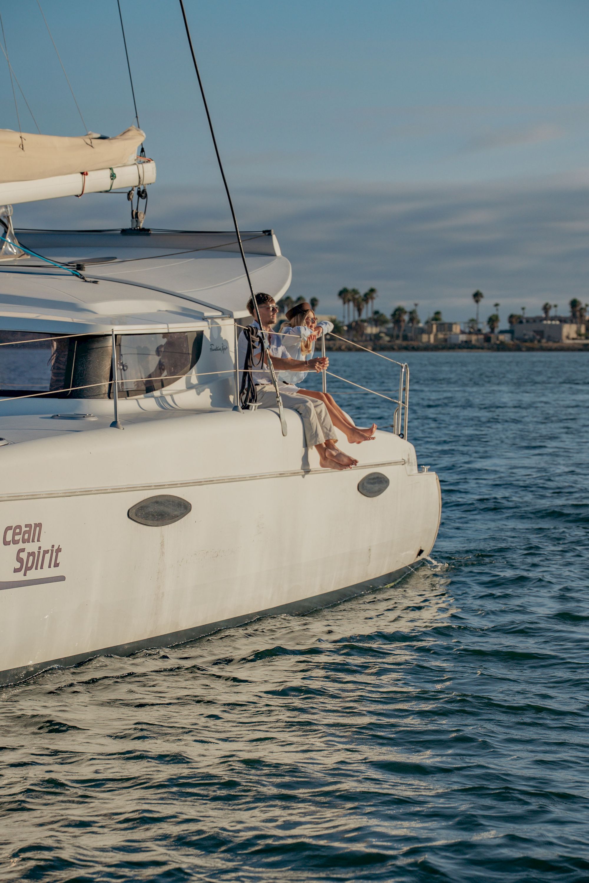 Two people relax on a white sailboat, sails up, cruising over blue water near a palm-lined coastline at sunset.