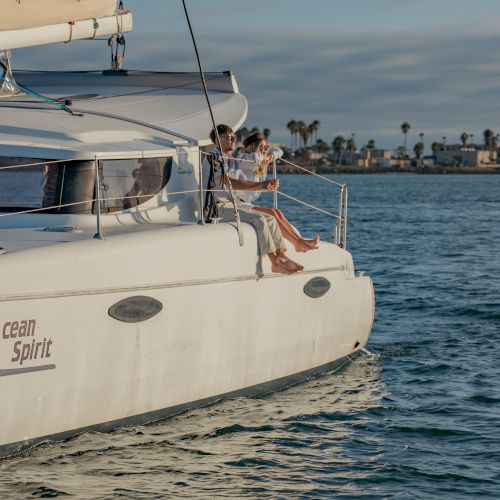 A small sailboat on open water with a couple lounging at the helm as palm-lined shore sits in the distance.