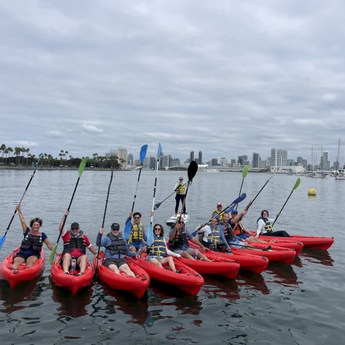 Kayakers in red boats paddle on calm water with palms and a downtown skyline under cloudy skies.
