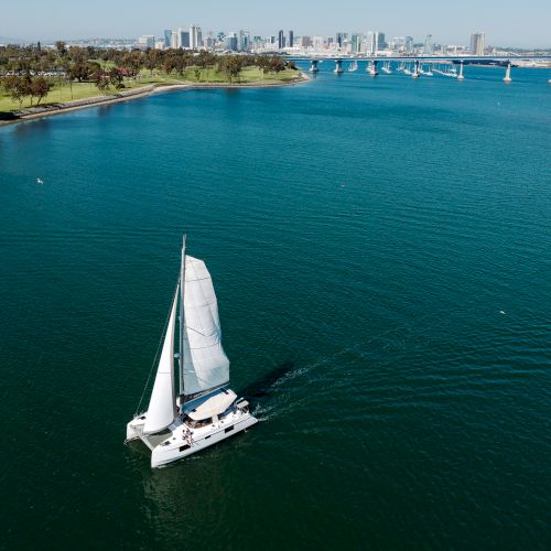 An aerial view of a sailboat on blue water, with a long bridge and a distant city skyline along a green shore.