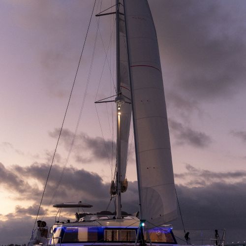 A sailboat/yacht with a tall mast and sails lit at dusk, tied on calm water under cloudy skies, with people onboard.
