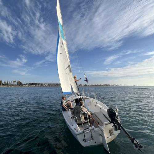 Sailboat glides on choppy blue water under billowing white-and-blue sails, with crew handling lines on a sunny day.
