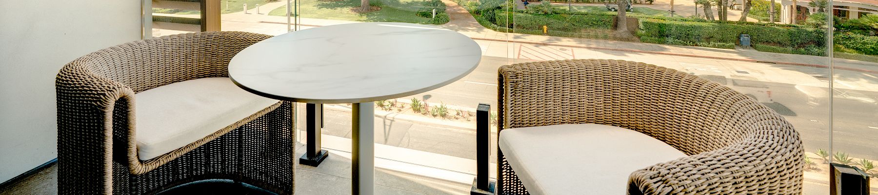Balcony patio with wicker seating, a round table, and a sunny resort courtyard of trees, palms, and white buildings beneath blue sky.