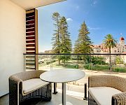 Balcony patio with wicker seating, a round table, and a sunny resort courtyard of trees, palms, and white buildings beneath blue sky.