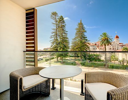 Balcony patio with wicker seating, a round table, and a sunny resort courtyard of trees, palms, and white buildings beneath blue sky.
