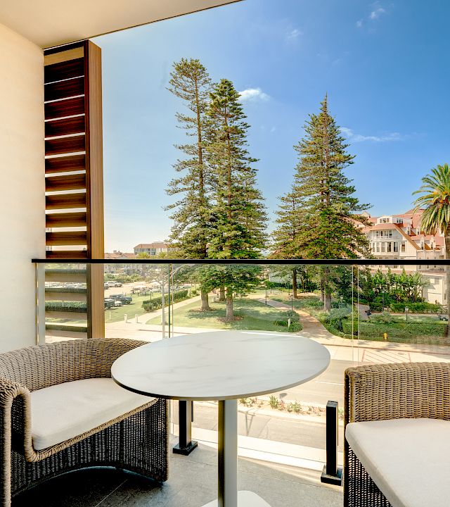 Balcony patio with wicker seating, a round table, and a sunny resort courtyard of trees, palms, and white buildings beneath blue sky.