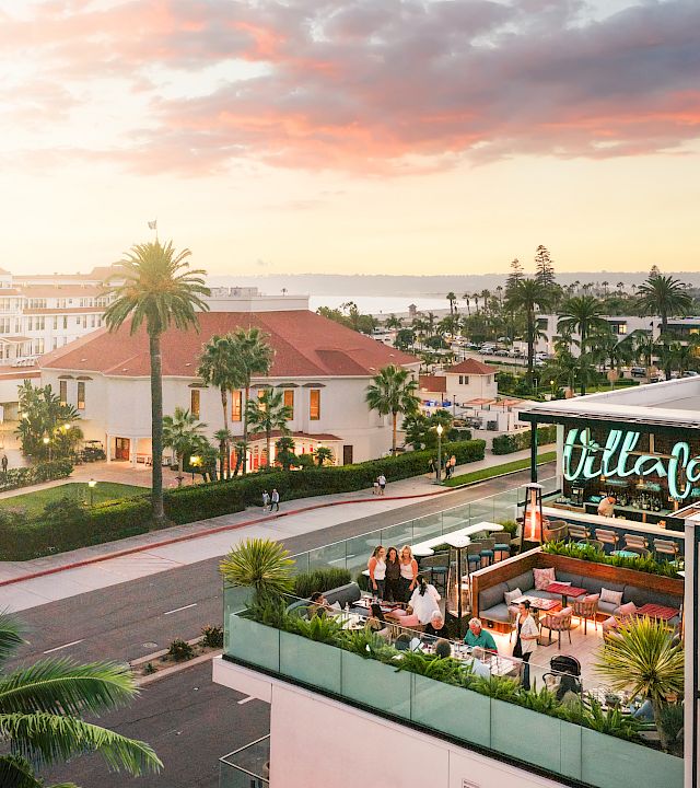 A sunny resort courtyard with palm trees, white buildings, and a rooftop terrace bar glowing at sunset in warm coastal light.