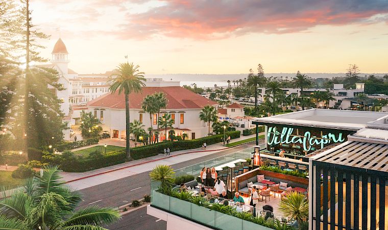 A sunny resort courtyard with palm trees, white buildings, and a rooftop terrace bar glowing at sunset in warm coastal light.