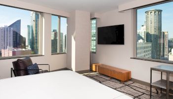 A modern hotel room with a bed, TV, armchair, and large windows offering a cityscape view, featuring tall buildings and clear skies.