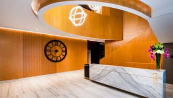 A modern lobby features a marble counter, wooden walls, a large clock, spiral ceiling, and decorative lighting with flowers on the desk.