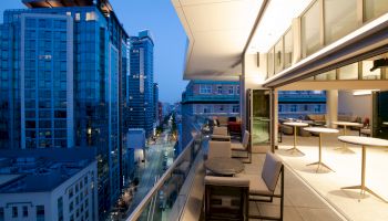 A modern rooftop patio at dusk, with tables and chairs, overlooks a city street lined with tall buildings.