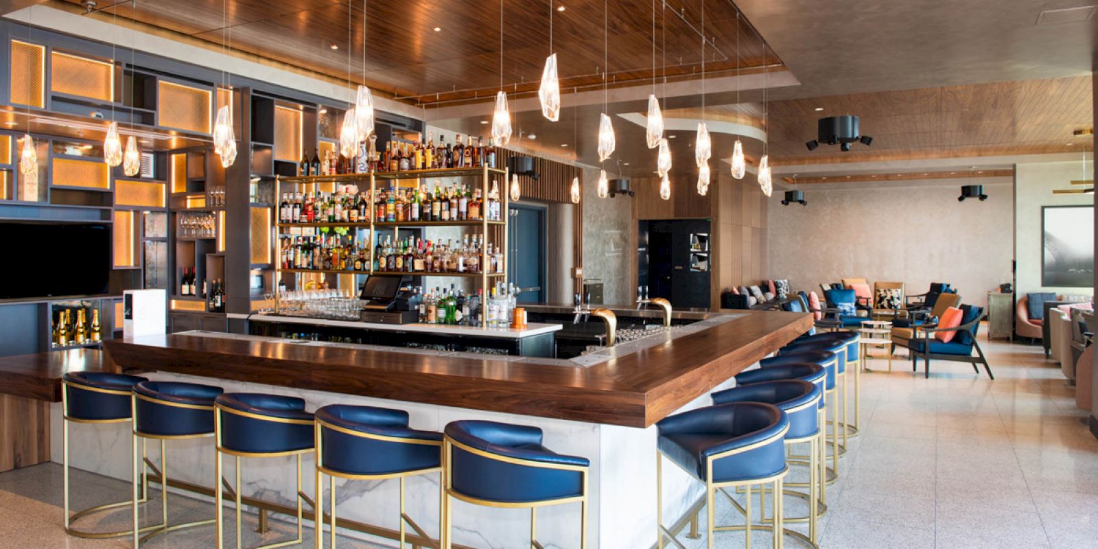 A modern, stylish bar with a long counter, blue chairs, hanging lights, and a well-stocked shelf of bottles, in a spacious lounge area.