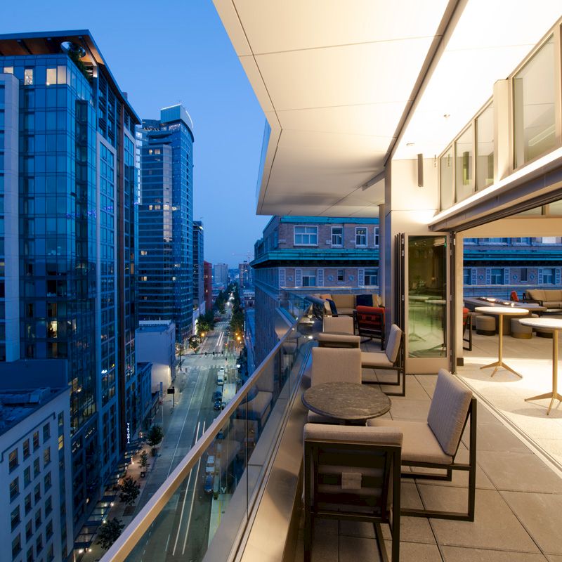 A modern, open balcony with seating overlooks a city street lined with tall buildings at dusk, lights illuminating the scene.