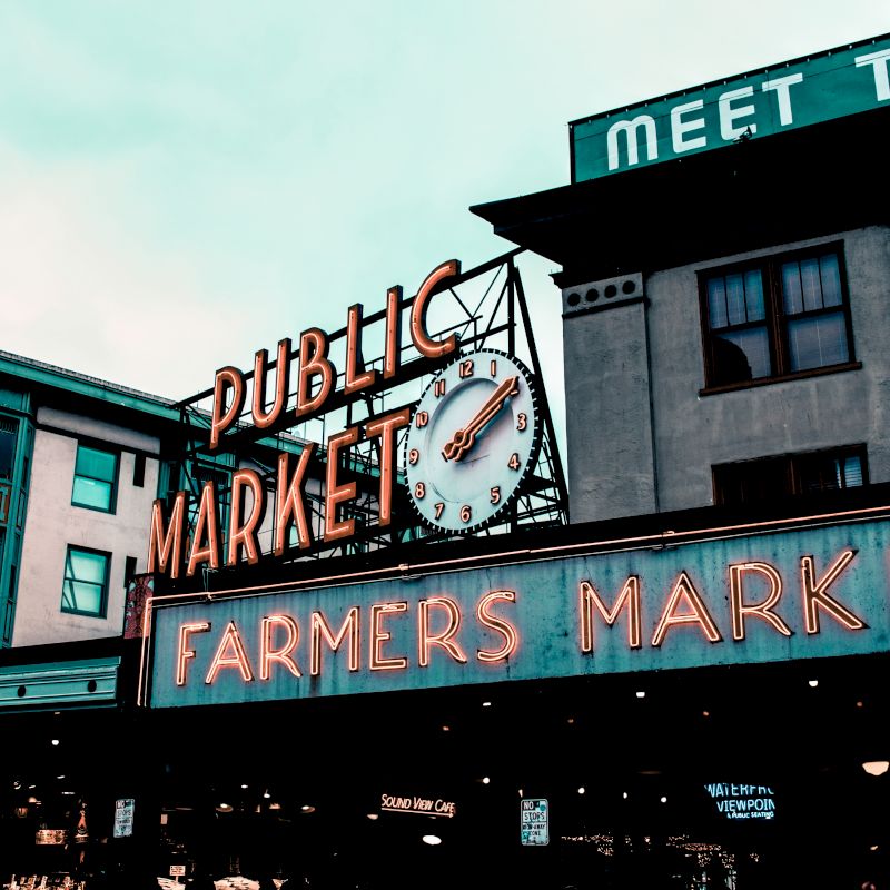 The image shows the entrance to a public farmers market with a large clock and neon signs on urban buildings.