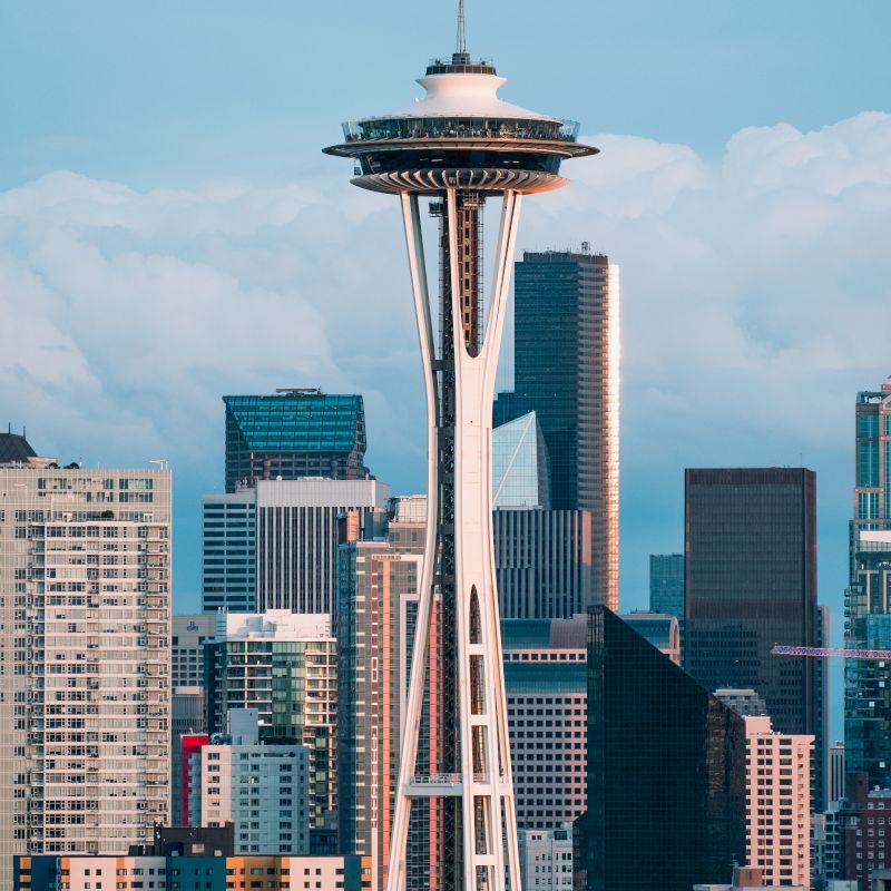 The image shows the Space Needle in Seattle, surrounded by modern skyscrapers and buildings against a cloudy sky backdrop.