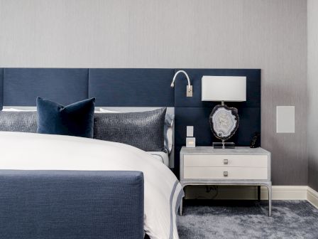A modern bedroom with a blue upholstered bed, gray pillows, and a nightstand with a lamp and decorative item, styled in a sleek design.