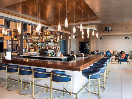 The image shows a modern bar with blue stools, a central counter, hanging lights, and shelves stocked with bottles, in a stylish lounge.