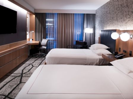 The image shows a modern hotel room with two beds, a TV, a desk with a chair, and stylish decor, including patterned wallpaper.