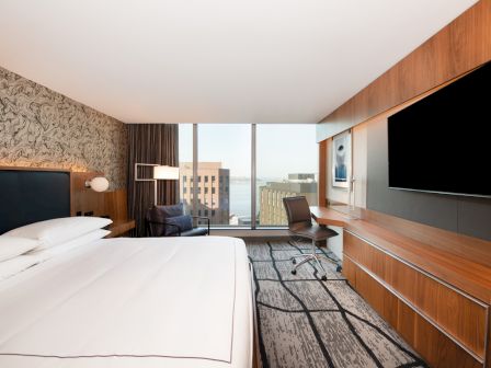 The image shows a modern hotel room with a large bed, a wall-mounted TV, a desk with a chair, and a window offering a city view.