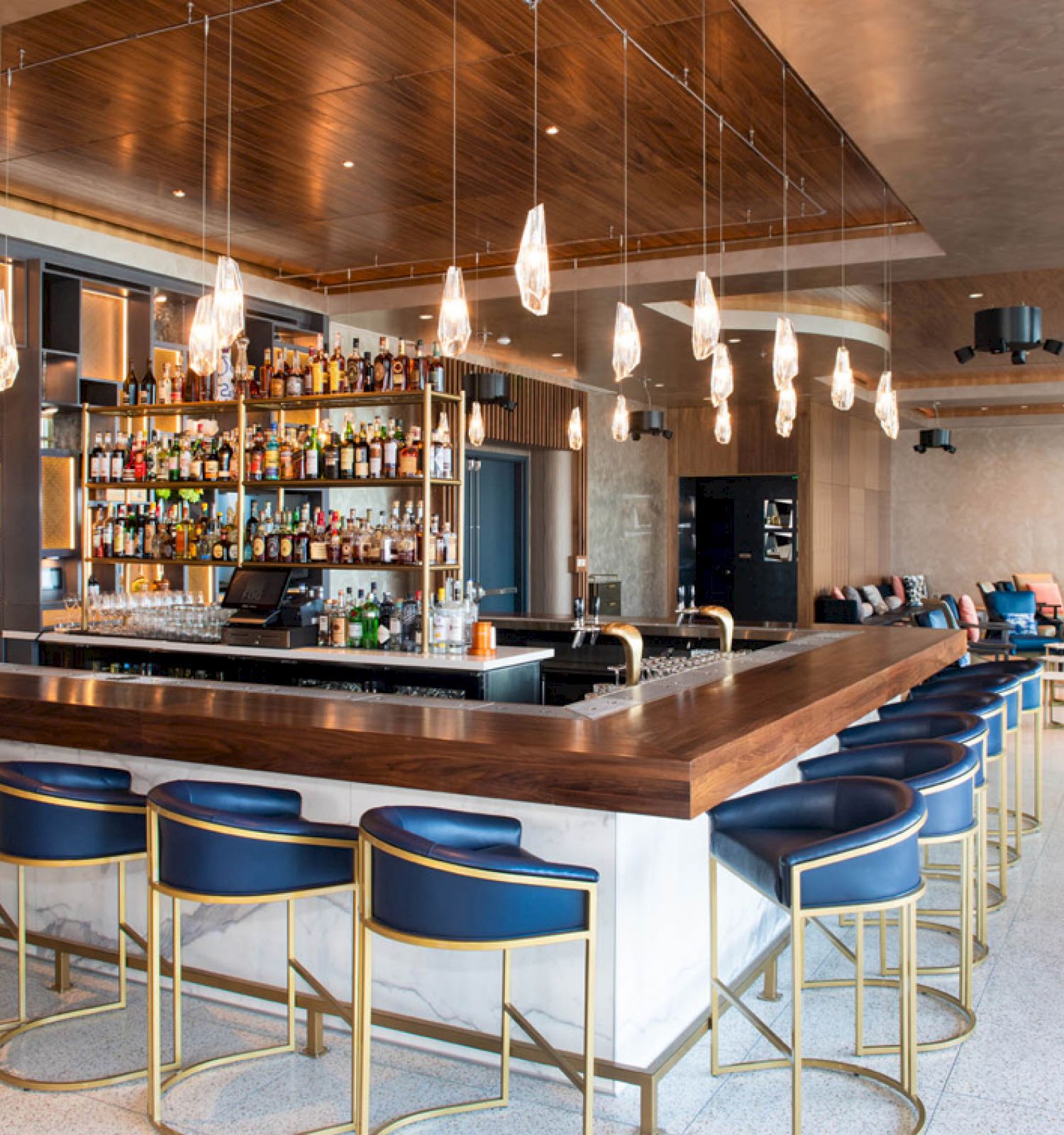 A modern bar with a wooden countertop, blue cushioned stools, hanging lights, and a fully stocked shelf of spirits.